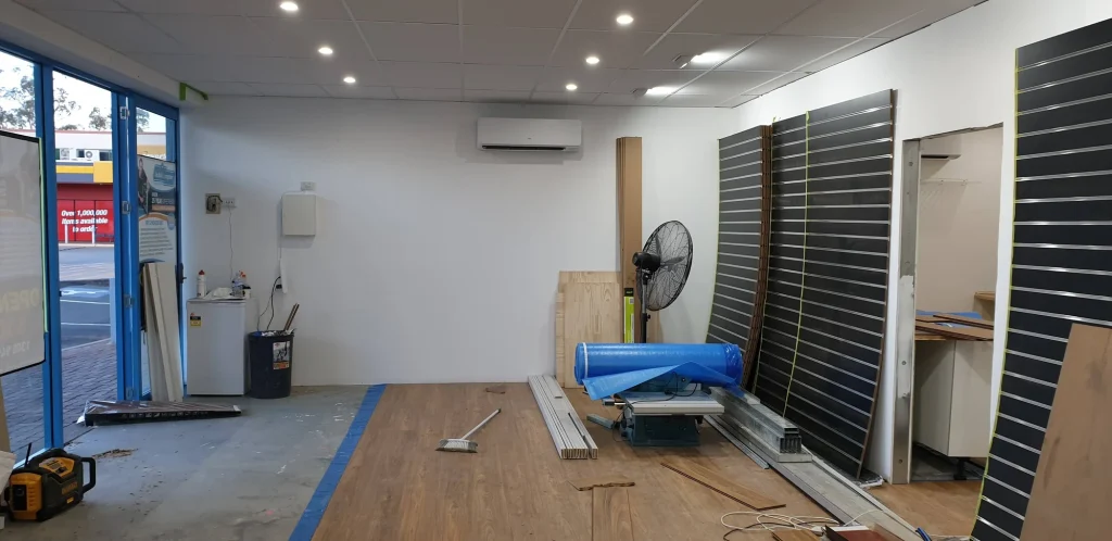 Partly installed timber-look flooring beneath a completed ceiling, with black display panels stacked against the wall