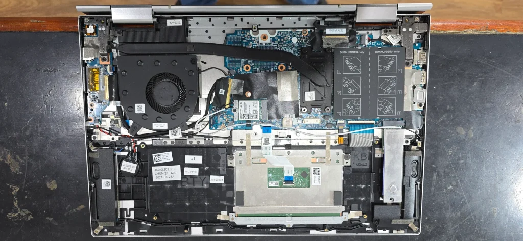 Laptop on a workshop bench with its bottom cover removed, showing a cooling fan, heat pipe, motherboard and memory slots